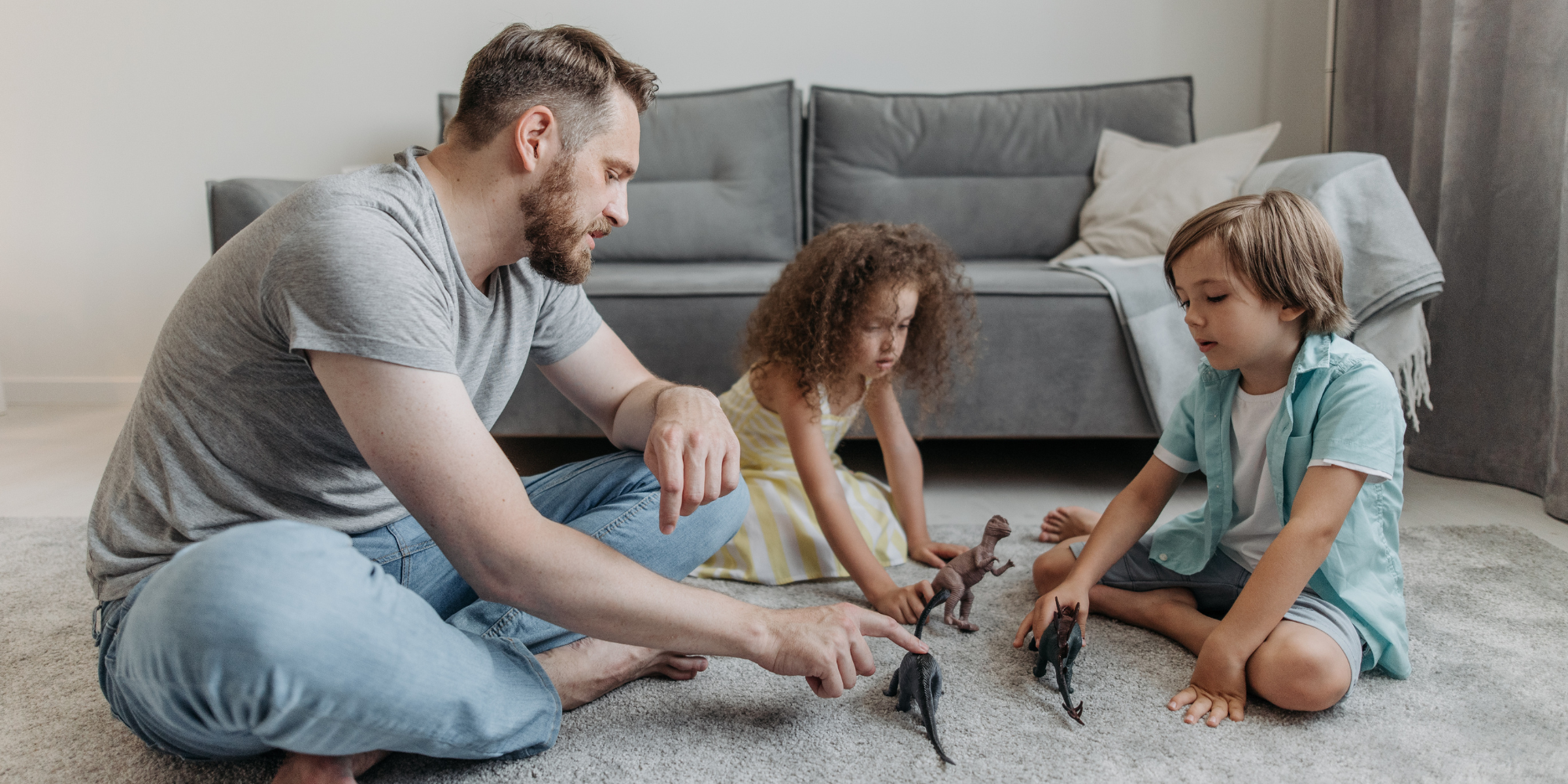 Un père joue au sol avec ses deux jeunes enfants, un garçon et une fille, en faisant marcher des figurines de dinosaures dans le salon.